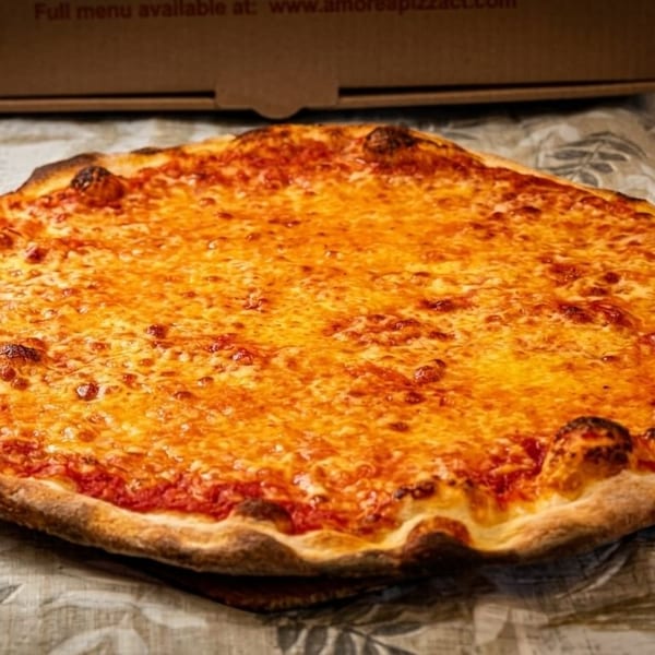A large, round cheese pizza with a golden-brown crust sits on a patterned tablecloth.