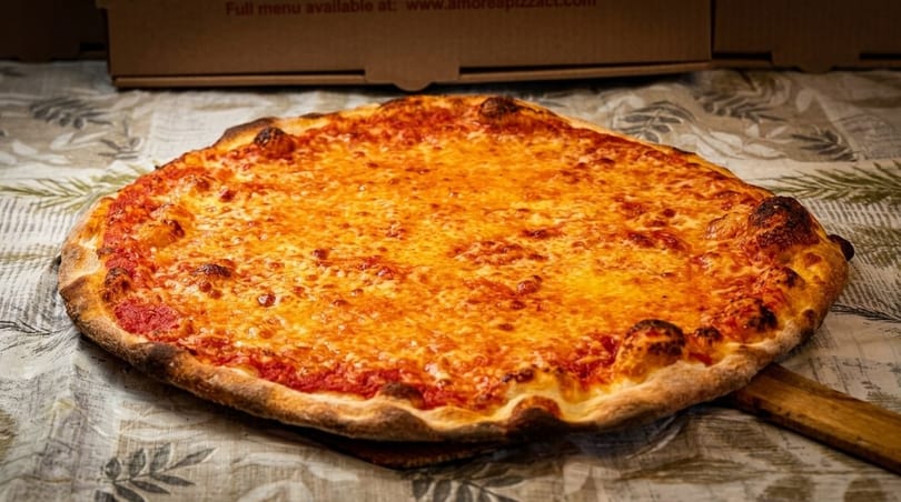 A large, round cheese pizza with a golden-brown crust sits on a patterned tablecloth.